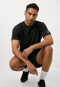 Schwarzes T-Shirt mit kurzen Ärmeln, das ein Logo auf der linken Brust und ein strukturiertes Design zeigt, kombiniert mit dunklen Shorts und schwarzen Turnschuhen.