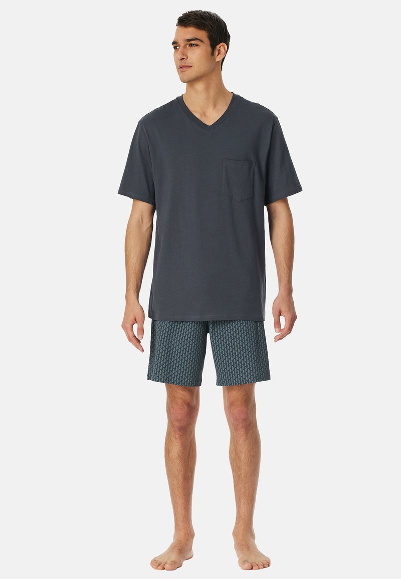 Dunkelgraues Baumwoll-V-Ausschnitt-T-Shirt mit einer Tasche, kombiniert mit gemusterten türkisfarbenen Shorts mit geometrischem Design. Barfuß gehalten.
