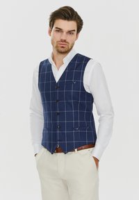 Granatowa kamizelka w kratę o dopasowanym kroju, wykonana z teksturowanej tkaniny. Posiada pięć guzików oraz dwie kieszenie z przodu, noszona na białej koszuli.