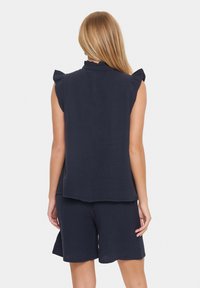 Tumši zila bezpiedurkņu bluse ar ruffled plecu akcentiem un teksturētu audumu, apvienota ar atbilstošām šortiem, lai radītu saskaņotu izskatu.