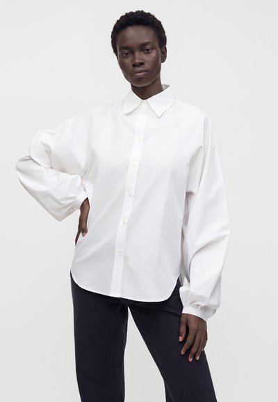 Personne portant une chemise blanche ample à boutons avec des manches surdimensionnées et un pantalon noir, debout avec une main sur la hanche devant un fond uni.