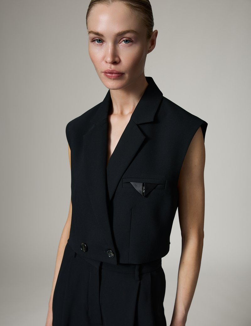Woman wearing a sleeveless black blazer with two buttons and a small pocket detail, standing against a plain light gray background.