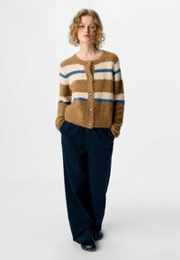 Person mit lockigem blonden Haar, der eine braun- und cremefarbige gestreifte flauschige Strickjacke, eine marineblaue weit geschnittene Hose und schwarze Clogs trägt, steht mit den Händen in den Taschen.