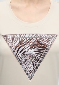 Μπεζ πουκάμισο με μεγάλο τριγωνικό λογότυπο Guess με ρίγες ζέβρας, στρας και κείμενο "U.S.A. Washed Jeans" στο στήθος.