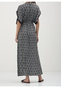 Robe maxi en tissu léger avec un motif de feuilles noir et blanc, taille cintrée et larges manches. Sandales noires plates aux pieds.