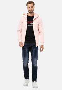Lichtroze zip-up hoodie met een trekkoord capuchon, gecombineerd met een zwarte graphic t-shirt en donkerdenim jeans. Witte sneakers maken de outfit compleet.