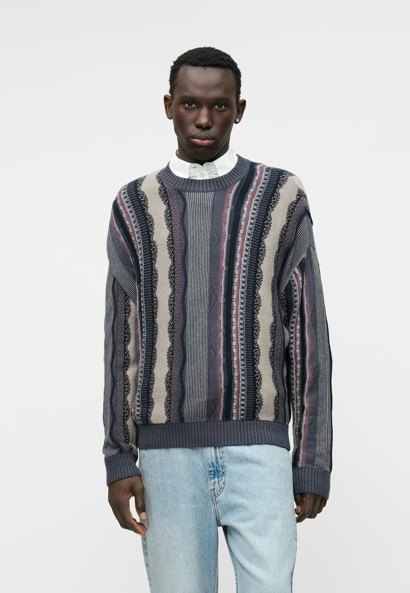 Πλεκτό πουλόβερ με συνδυασμό γκρι, ναυτικού και κρεμ ρίγες με υΦές. Ραμμένα μανίκια και τελείωμα, στρογγυλή λαιμόκοψη πάνω από ένα λευκό πουκάμισο με γιακά.