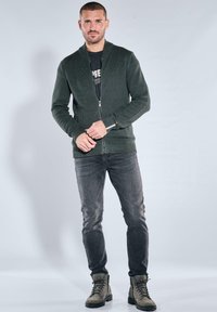 Dunkelgrüner, strukturierter Reißverschluss-Cardigan mit Schalkragen, getragen über einem schwarzen T-Shirt und kombiniert mit grauen Slim-Fit-Jeans und beigen Stiefeln.