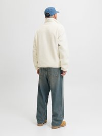 Fleecasta jakna u off-white boji s visokim ovratnikom, u kombinaciji s širokim plavim trapericama i bež obućom, prikazana sa stražnje strane.