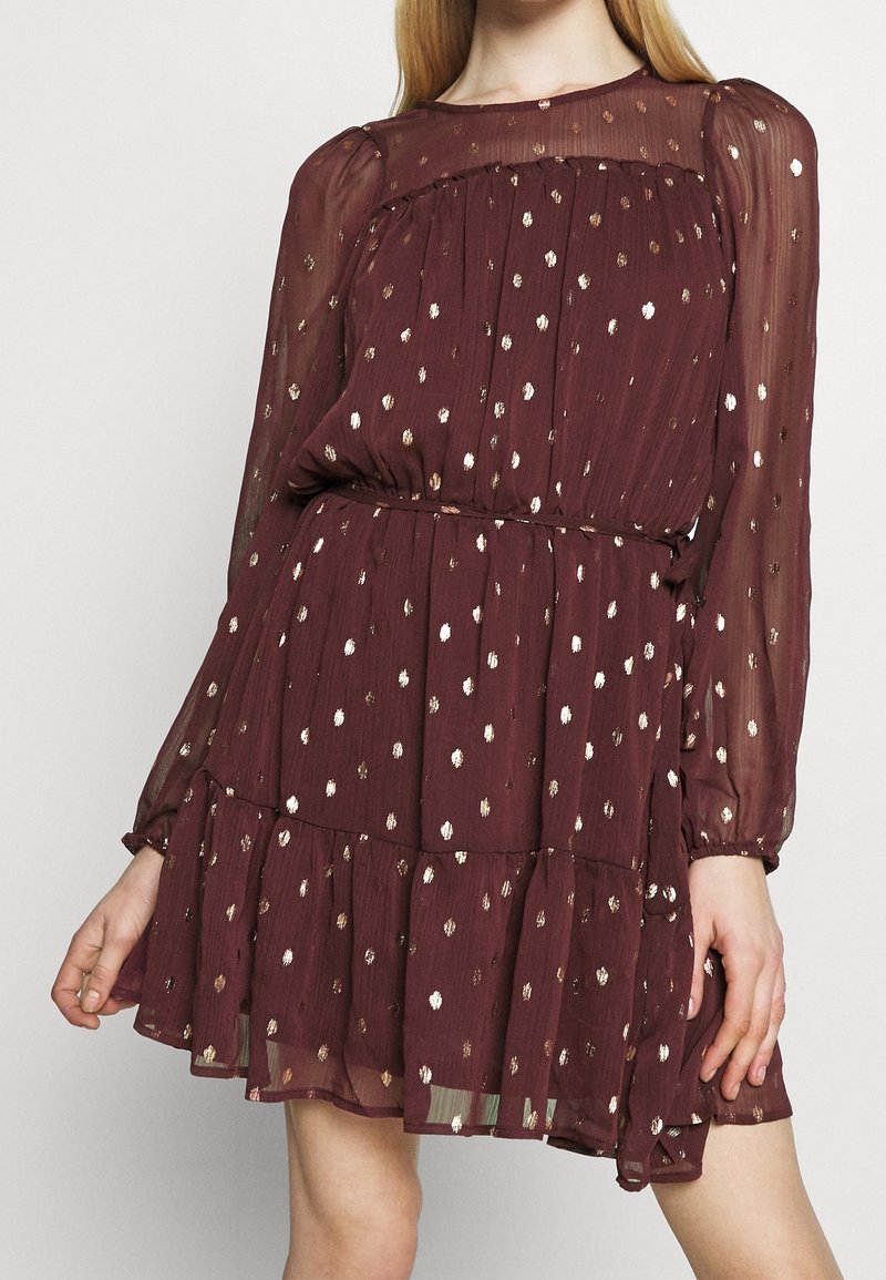 Burgundy klänning i genomskinligt tyg med långa ärmar, prydd med guld- och vitprickiga mönster samt en lager-på-lager volangkant.