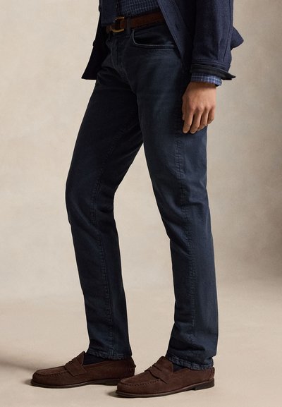 Vyras dėvi tamsiai mėlynas džinsines kelnes, rudos spalvos zomšines loaferius, mėlyną languotą marškinių ir tamsiai mėlyną švarką, stovintis ant smėlio spalvos fono.