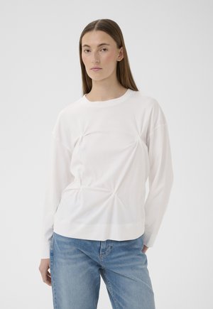 Žena s dlhými hnedými vlasmi, oblečená v bielom dlhom rukáve s nariasenými detailmi a modrých džínsoch, stojaca pred jednoduchým svetlým pozadím.