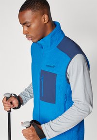 Blue fleece vest with a high collar, gray long sleeves, and navy blue chest pocket. Features zippered closures and is paired with trekking poles.