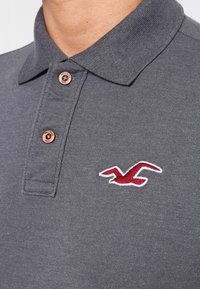 Šedé polo tričko se strukturovaným límcem, vybavené třemi hnědými knoflíky a červeným vyšitým logem na levé části hrudi.