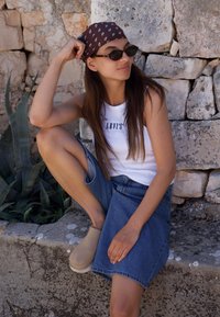 Balta maikė be rankovių su "LEVI'S" tekstu, mėlynos džinsinio audinio sijonas, rudas šalikas ir saulės akinių. Šviesiai rudos spalvos šlepetės, sėdi prieš akmeninę sieną apsupta žalumos.