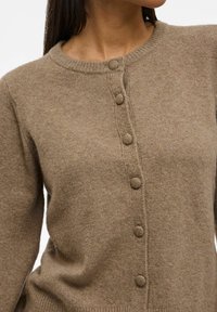 Femme portant un cardigan en tricot marron avec six boutons recouverts de tissu et un col rond, montrée de la nuque à la taille.