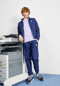 Ναυτικό Nike tracksuit από ελαφρύ ύφασμα, που περιλαμβάνει σακάκι με φερμουάρ και ταιριαστά παντελόνια. Μαύρα και μπλε αθλητικά παπούτσια ολοκληρώνουν την εμφάνιση.