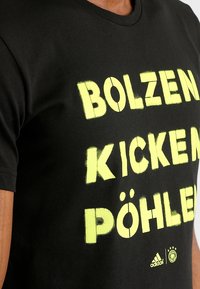 Camiseta de algodón negra con texto en amarillo spray pintado "BOLZEN KICKEN PÖHLEN" y logo de Adidas con un emblema redondo. Mangas cortas.