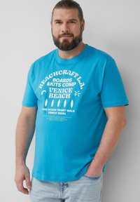 Turquoise katoenen t-shirt met witte tekst en graphics. Korte mouwen, ronde halslijn, ontspannen pasvorm. Combinatie met lichte denim jeans.