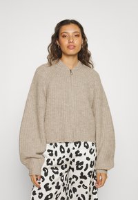 Lichtbruin, geribbeld zip-up sweater met oversized, ballonmouwen en een hoge kraag, gecombineerd met zwart-witte gestreepte broek.