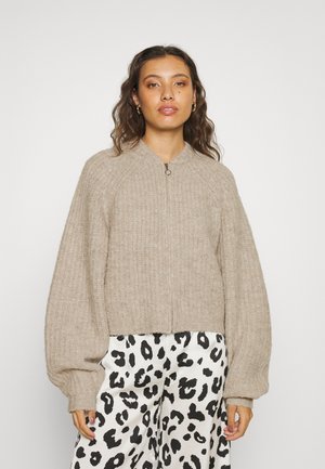 Hellbrauner, gerippter Zip-Up-Pullover mit übergroßen Ballonärmeln und hohen Kragen, kombiniert mit gemusterten schwarz-weißen Hosen.