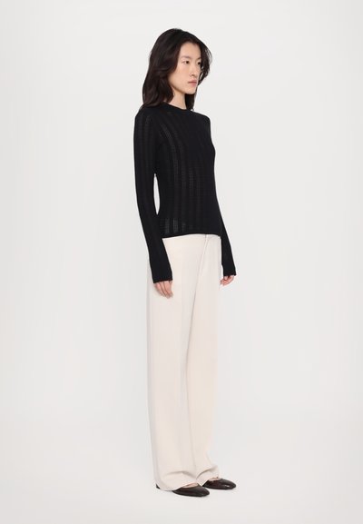 Sort strikket sweater med lange ærmer og et struktureret mønster, parret med beige bredbenede bukser og mørke sko.