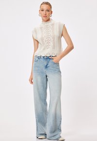 Knit sleeveless top in cream with a lace pattern, paired with light blue wide-leg jeans. Smooth texture with scalloped hem.