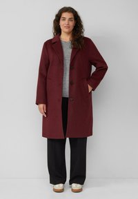 Vrouw in een donkerrode, knopenjasje, grijze trui, zwarte broek en witte sneakers, staande tegen een effen lichte achtergrond.