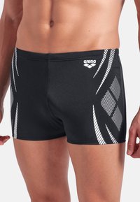 Svarta badshorts i elastiskt material, med vita grafiska mönster och detaljer längs sidorna. Logotyp placerad på midjebandet.