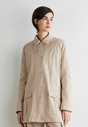 Frau mit kurzen dunklen Haaren, die einen beigen, mit Knöpfen versehenen Mantel mit Klappentaschen trägt, steht vor einer schlichten hellen Wand und schaut nach rechts.