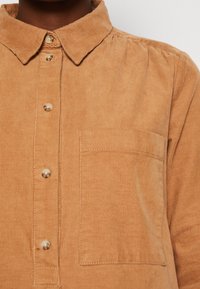 Beige Cordhemd mit Kragen, ausgestattet mit Knöpfen und einer Brusttasche. Strukturierter Stoff mit vertikalen Rillen.