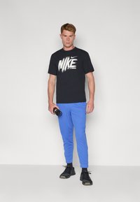 Camiseta negra de Nike con logo blanco, combinada con pantalones ajustados de color azul brillante. El modelo sostiene una cámara y lleva zapatillas deportivas negras.