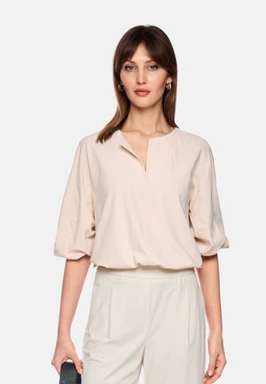 Woman with straight brown hair wearing gold hoop earrings, a beige loose blouse with puffed sleeves, and light-colored high-waist pants.