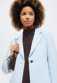 Femme aux cheveux bouclés portant un blazer bleu clair sur un chemisier noir à pois, tenant un petit sac à main sombre avec une chaîne argentée.