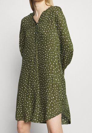 Woman wearing loose-fitting, olive green dress with white irregular dots, long sleeves, and a band collar.