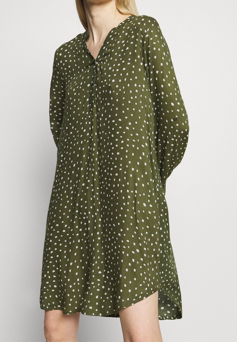 Femme portant une robe ample vert olive avec des points blancs irréguliers, manches longues et col montant.