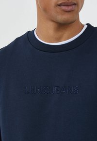 Hombre vestido con sudadera azul marino con "LIU·JO JEANS" bordado en el pecho sobre una camiseta de cuello redondo blanca, recortada a la altura de la boca y los hombros.