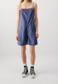 Sininen raidallinen farkkuhaalari, jossa on spagettiolkaimet, neliöpääntie ja nauhakoristeet; sisältää joustavan vyötärön ja löysät shortsit.