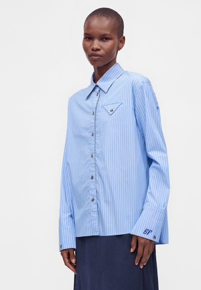 Personne portant une chemise à rayures bleu et blanc avec des boutons et un col pointu, ainsi qu'une jupe bleu marine, debout devant un fond clair uni.