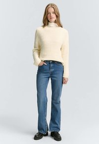 Krämfärgad stickad tröja med ett strukturerat mönster, kombinerad med blåa denimflare-jeans och svarta platåskor. Enkel, figurnära design.