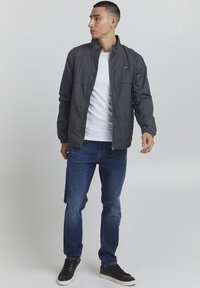 Grå letvægtsjakke med høj krave, frontlynlås og elastiske manchetter, parret med blå denimjeans og sorte sneakers.