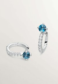 Los pendientes de aro de plata presentan una gema azul engastada en un soporte de estilo garra, con piedras transparentes a lo largo del aro para un detalle adicional.