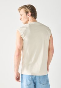 Light beige sleeveless t-shirt with a relaxed fit, made of soft fabric. Features a simple neck design and clean seams, paired with blue denim shorts.