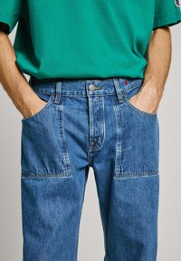 Jeans de denim azul con un corte relajado, que cuentan con bolsillos laterales y un acento rojo visible cerca de la cintura. Combinados con una camiseta verde.