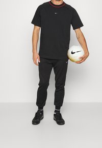 Muž v černém sportovním oblečení Nike, který drží bílý a zlatý fotbal Nike v pravé ruce, stojí na bílém pozadí.