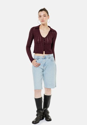 Femme debout, portant un cardigan bordeaux en tricot boutonné, un short en jean clair à hauteur du genou, des bottes noires montant jusqu'aux genoux et de grandes boucles d'oreilles créoles.