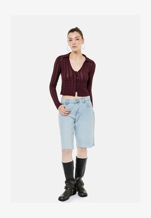Femme debout, portant un cardigan bordeaux en tricot boutonné, un short en jean clair à hauteur du genou, des bottes noires montant jusqu'aux genoux et de grandes boucles d'oreilles créoles.