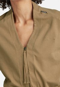 Khaki zip-up kombinéza s výstřihem do V, vyrobená z měkké látky; obsahuje malou značkovou nálepku a texturované táhlo zipu.