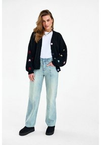 Woman wearing a black cardigan with colorful star patterns, white shirt, light blue wide-leg jeans, and black platform shoes, standing with one hand in pocket.