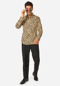 Camicia con stampa leopardata in beige, con accenti arancioni e neri; abbinata a pantaloni e scarpe nere; persona che aggiusta occhiali da sole neri.